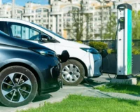 Two electric cars charging at a public charging station in an urban setting, with buildings in the background and green grass in the foreground.