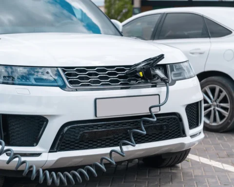 Close-up of a white hybrid SUV plugged into a charging station, showcasing its front grille and charging cable connected to the port, parked on a paved lot next to another vehicle