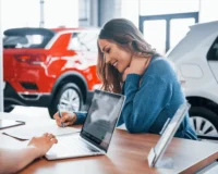 Smiling woman signing car purchase documents at a dealership desk with laptops and new vehicles in the background.