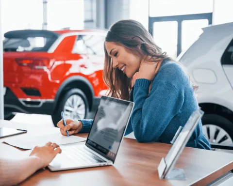 Smiling woman signing car purchase documents at a dealership desk with laptops and new vehicles in the background.