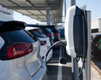 Electric vehicles parked in a row at a charging station under a solar-covered parking area, with one EV charger in the foreground.