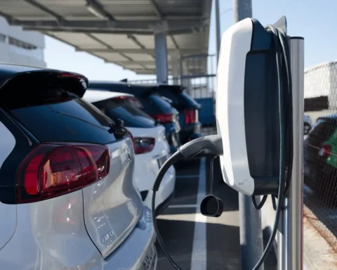 Electric vehicles parked in a row at a charging station under a solar-covered parking area, with one EV charger in the foreground.