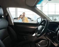 A smiling couple stands in a car dealership, viewed from inside a sedan through the driver’s side window. They are shopping for the perfect family-friendly sedan.
