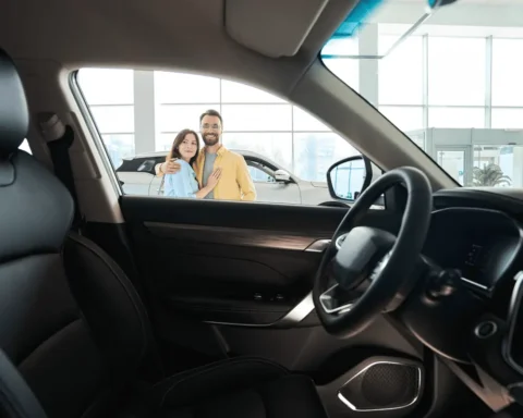A smiling couple stands in a car dealership, viewed from inside a sedan through the driver’s side window. They are shopping for the perfect family-friendly sedan.