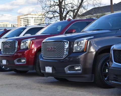 A row of GMC SUVs parked in a dealership lot, featuring various colors including red, white, and dark gray, with a focus on the large front grilles and GMC badges.