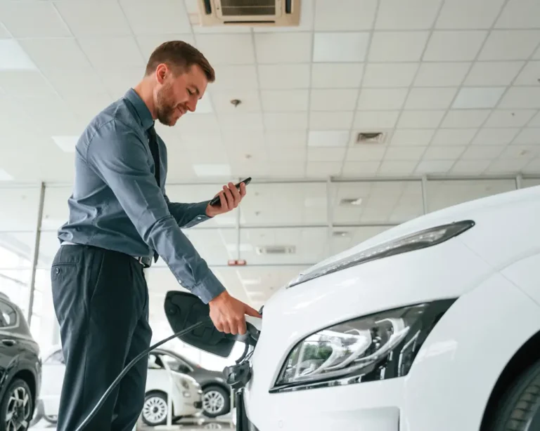 A man inspects a used white electric vehicle inside a dealership, holding a charging cable in one hand and a smartphone in the other, checking battery stats or service history before buying a used EV.