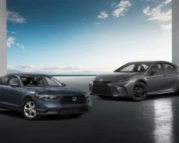 Two midsize hybrid sedans— a blue 2025 Honda Accord Hybrid (left) and a grey 2025 Toyota Camry Hybrid (right)—parked side by side in front of a coastal backdrop with a clear blue sky and modern architectural columns.