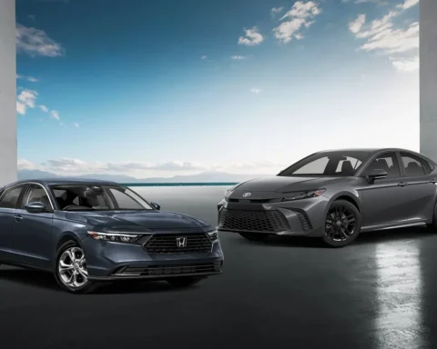 Two midsize hybrid sedans— a blue 2025 Honda Accord Hybrid (left) and a grey 2025 Toyota Camry Hybrid (right)—parked side by side in front of a coastal backdrop with a clear blue sky and modern architectural columns.