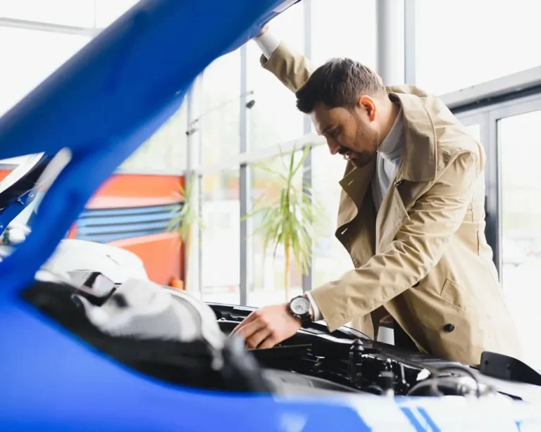 A man in a beige trench coat inspecting the engine of a blue car with the hood open inside a well-lit showroom or garage, possibly evaluating a used vehicle before purchase.