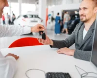 A smiling man in a grey blazer receiving car keys from a dealership representative across a white desk, symbolizing the completion of a car lease or purchase in a showroom environment.