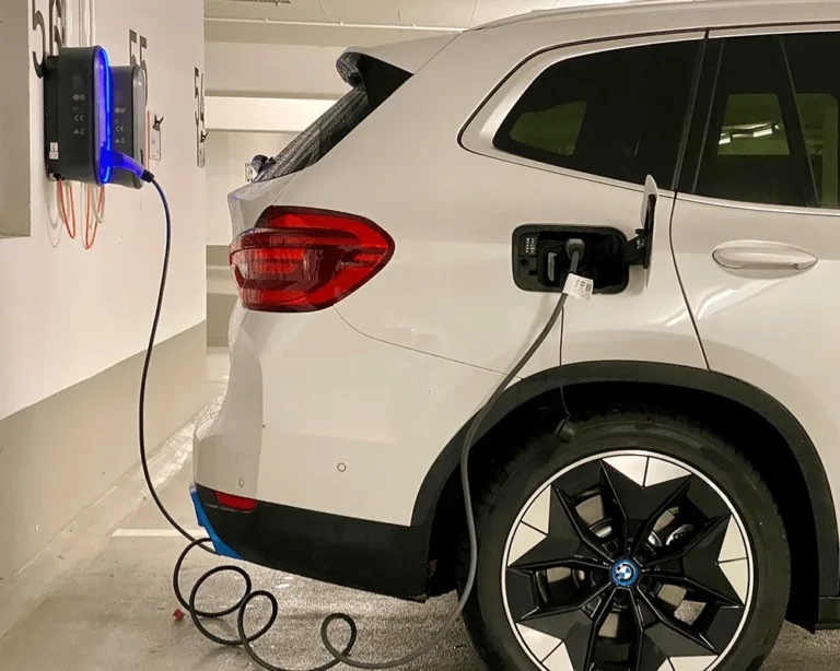 A white BMW electric vehicle is plugged into a wall-mounted charging station in an indoor parking garage, illustrating the process of charging an EV battery.